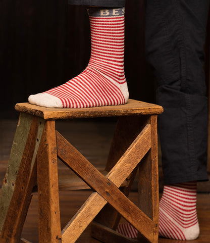 A person in dark pants stands on a wooden step stool, wearing Best Made Co. Standard Wool Socks—red and white striped merino wool with a blue band reading "BEST"—designed for all-season comfort.