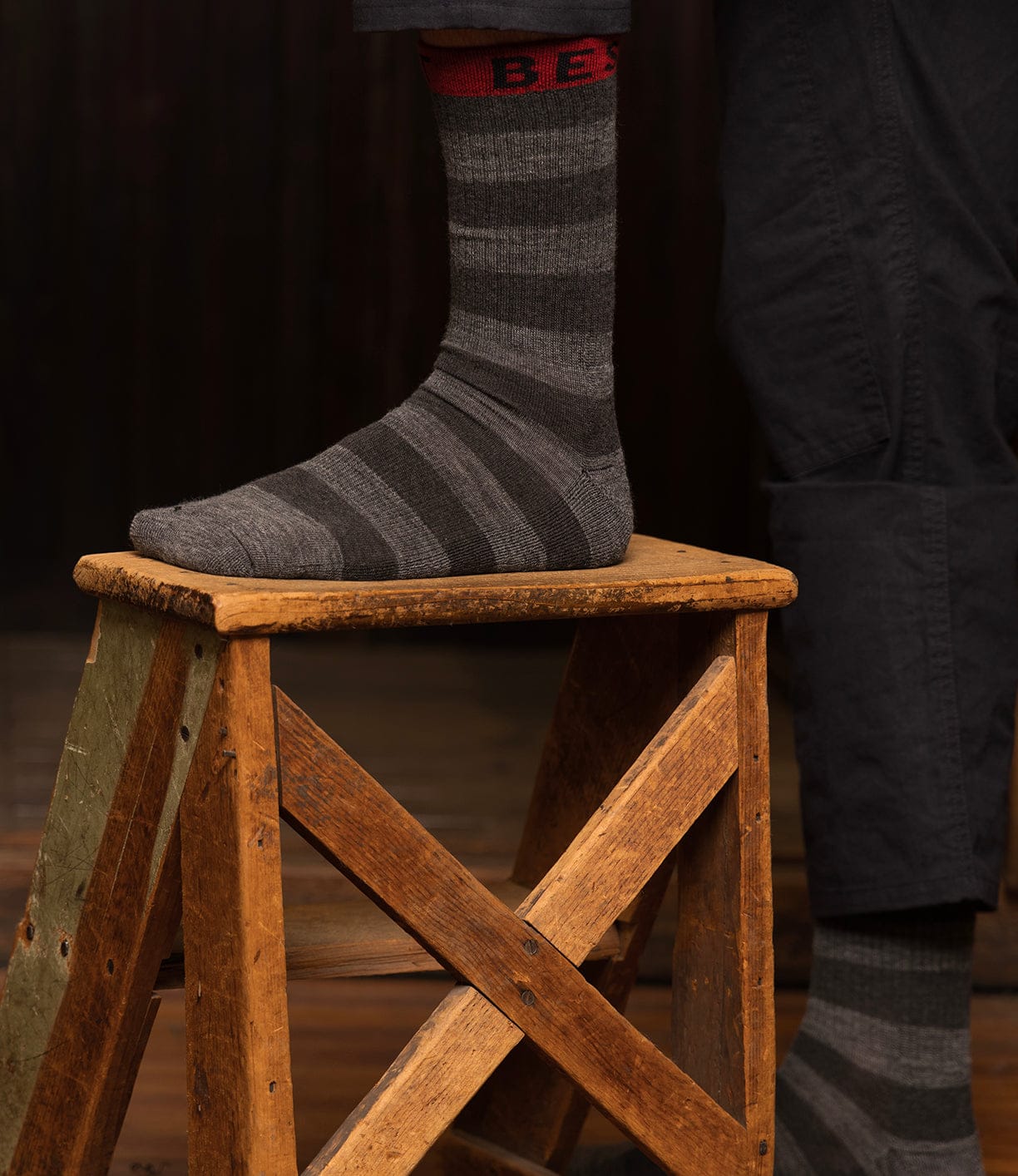 Wearing Best Made Co Standard Wool Socks in dark color, a person stands with one foot on a rustic wooden stool. Red lettering tops the moisture-wicking Merino wool socks, exuding cozy, all-season comfort against a dim backdrop.