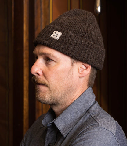 A man in a weather-resistant Shetland Cap of Courage by Best Made Co. and a blue button-up shirt stands in profile against a wooden background, arms crossed and looking forward.