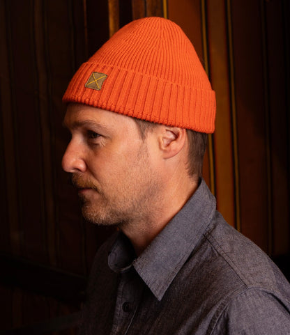 A man with light facial hair wears a bright orange Best Made Co. Cap of Courage and a gray button-up shirt, posing in profile against a dark, warmly lit background with vertical stripes.