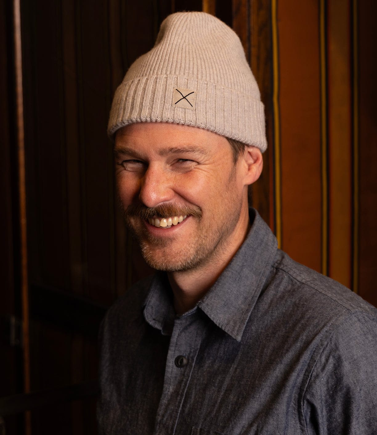 A man with a light beard and mustache smiles indoors, wearing the Best Made Co Cap of Courage lambswool cap and a blue button-up shirt, standing against a striped wooden background.