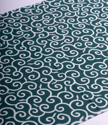 The Karakusa Tenugui by Best Made Co. is a hand-dyed Japanese cloth featuring a repeating white swirl on dark green in a diagonal pattern, inspired by wabi-sabi aesthetics.