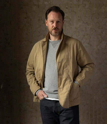 A man with short brown hair and a beard stands against a textured wall, wearing the Best Made Co. Possibilities Jacket Tan over a gray sweatshirt and dark jeans, with one hand in his jacket pocket.