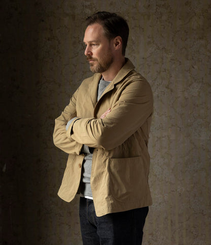 A man with light brown hair and a beard stands sideways, arms crossed, wearing the Best Made Co. Possibilities Jacket Tan over a gray shirt and dark jeans, against a textured, neutral wall.