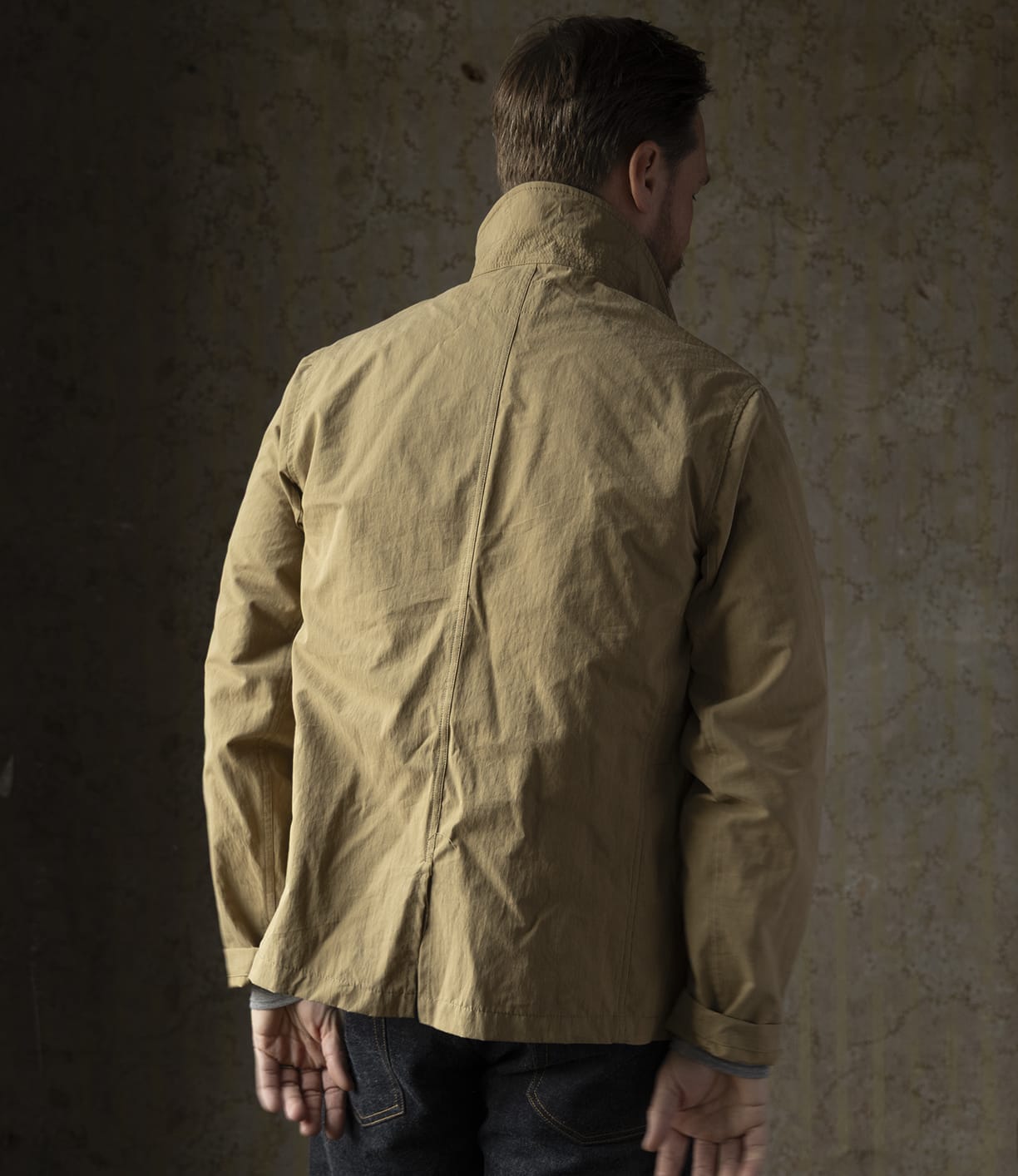 A person stands facing a textured wall, wearing Best Made Co.’s Possibilities Jacket Tan with the collar turned up and dark jeans. Their hands are visible behind their back.