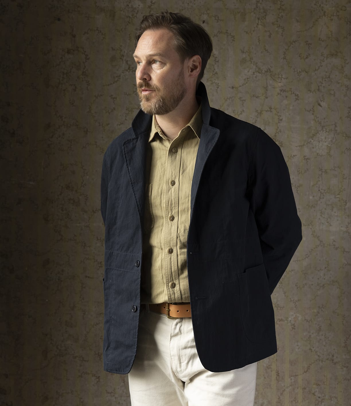 A man with light brown hair and a beard stands against a textured beige wall, wearing the Best Made Co. Possibilities Jacket in Navy over a tan button-up shirt, cream pants, and a brown belt, gazing thoughtfully to the side.