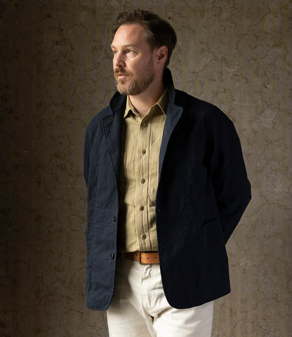 A man with light brown hair and a beard stands against a textured beige wall, wearing the Best Made Co. Possibilities Jacket in Navy over a tan button-up shirt, cream pants, and a brown belt, gazing thoughtfully to the side.