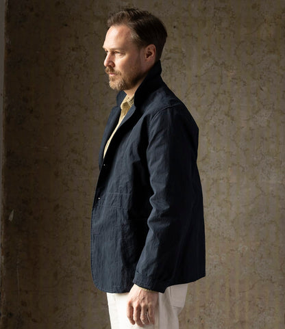 A man with light brown hair and a beard stands in profile against a textured neutral wall, wearing the Best Made Co. Possibilities Jacket Navy over a beige shirt and white pants.