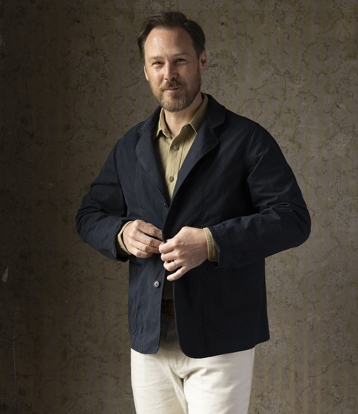 A man with light brown hair and a short beard stands against a textured wall, buttoning his Best Made Co. Possibilities Jacket Navy over a tan shirt and light-colored pants, smiling slightly.