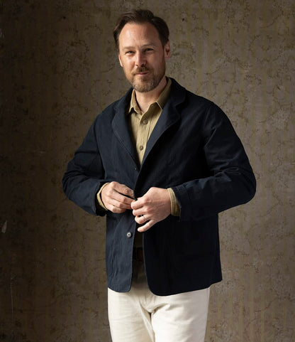 A man with light brown hair and a short beard stands against a textured wall, buttoning his Best Made Co. Possibilities Jacket Navy over a tan shirt and light-colored pants, smiling slightly.
