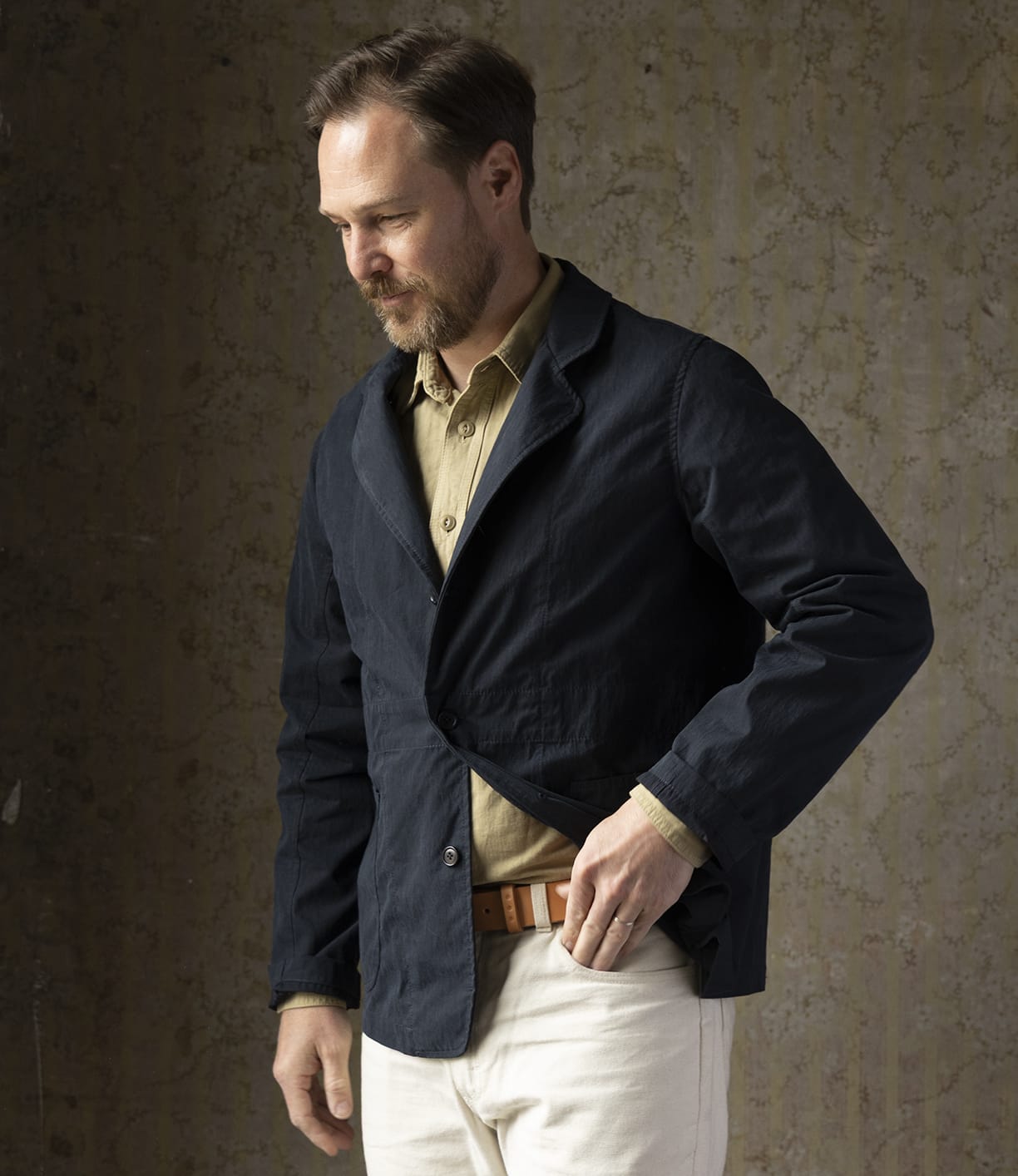 A man with short brown hair and a beard adjusts his Best Made Co. Possibilities Jacket Navy, layered under a tan cotton Cordura jacket and paired with white pants, standing before a textured neutral background.