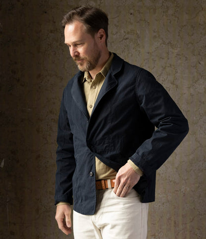 A man with short brown hair and a beard adjusts his Best Made Co. Possibilities Jacket Navy, layered under a tan cotton Cordura jacket and paired with white pants, standing before a textured neutral background.