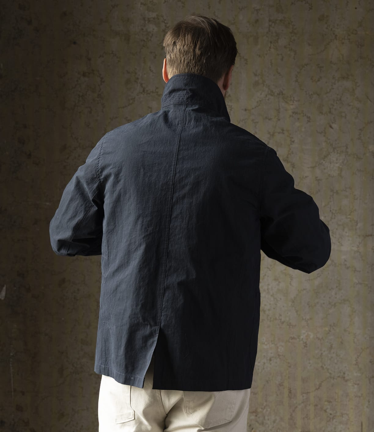 A person with short brown hair is seen from behind, wearing Best Made Co.'s Possibilities Jacket Navy and light-colored pants, standing against a textured, neutral-toned wall.