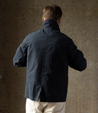 A person with short brown hair is seen from behind, wearing Best Made Co.'s Possibilities Jacket Navy and light-colored pants, standing against a textured, neutral-toned wall.