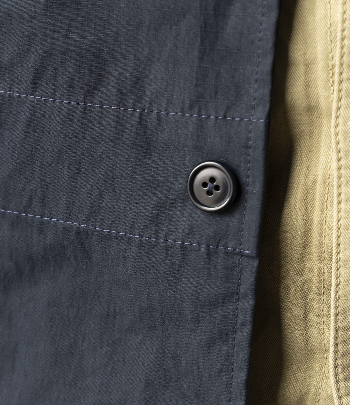 Close-up of the Possibilities Jacket Navy by Best Made Co., made from dark blue cotton Cordura with a black button and stitched seam, shown beside tan textured fabric.