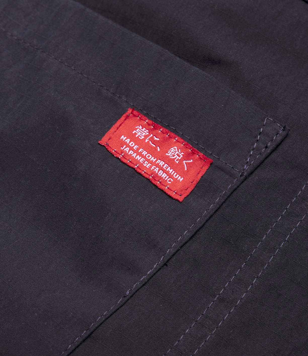 Close-up of navy ripstop fabric with stitched seams and a red label featuring Japanese characters and "MADE FROM PREMIUM JAPANESE FABRIC," on the lightweight Possibilities Jacket Navy by Best Made Co.