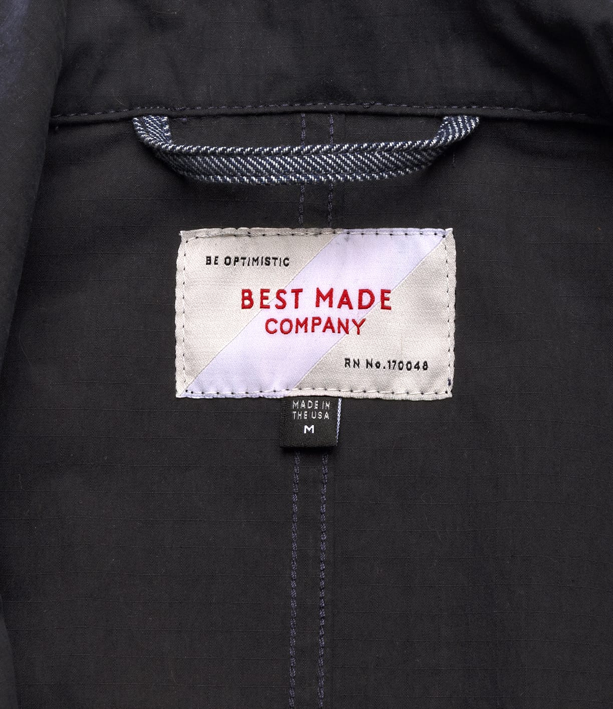 A close-up of the Possibilities Jacket Navy label shows "BE OPTIMISTIC" and "Best Made Co." in red, with size M and "MADE IN THE USA" printed on a smaller black tag below.