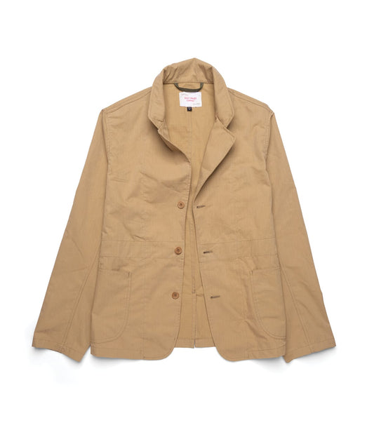 A man with short brown hair and a beard stands before a textured brown wall, wearing the Best Made Co Possibilities Jacket in Tan over a gray sweatshirt and organic cotton selvedge denim jeans, one hand in his pocket, the other at his waist.