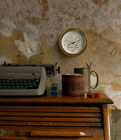 A vintage typewriter sits on a wooden desk beneath a Best Made Co. Weather Station, mounted on a textured wall. A mug of pencils, round tin, stapler, and red paper clips are also on the desk.
