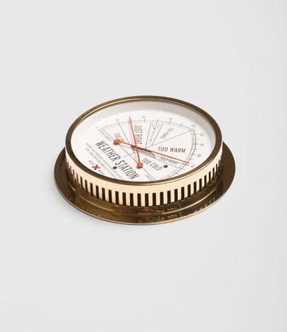 The Best Made Co. Weather Station is a round, vintage-style thermometer and hygrometer with a brass case and white dial; set on a white surface, its needle indicates the TOO WARM section for temperature and comfort levels.
