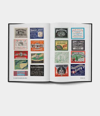 An open book, Best Made Co.’s Buchanan-Smith’s Axe Handbook (signed), displays colorful vintage axe and tool labels with unique designs, logos, and text arranged in a grid across two pages on a white background.