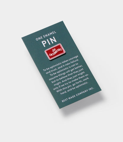 A rectangular green card displays a red enamel “Be Optimistic” pin. Beneath, white text celebrates persistence and optimism—key for wilderness survival. At the bottom, the card features Best Made Co., maker of the Be Optimistic Pin.