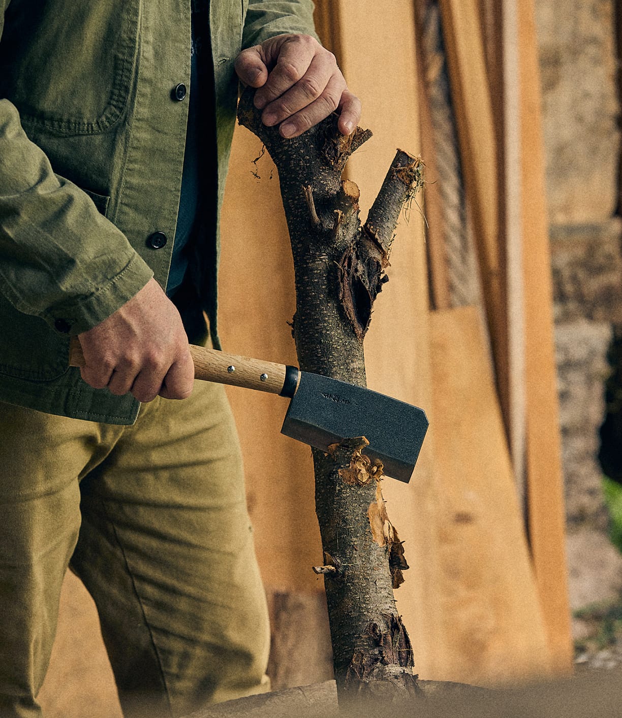 Wearing a green jacket, a person uses the Best Made Co. Azuma Hatchet to chop bark from a branch, with wood panels in the background.