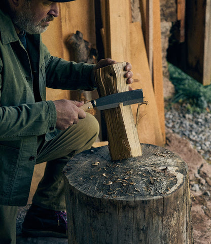 Wearing an olive jacket, a person uses the Best Made Co. Sayanata Hatchet—a large carbon steel blade—to split a piece of wood atop a tree stump, surrounded by lumber and gravel.