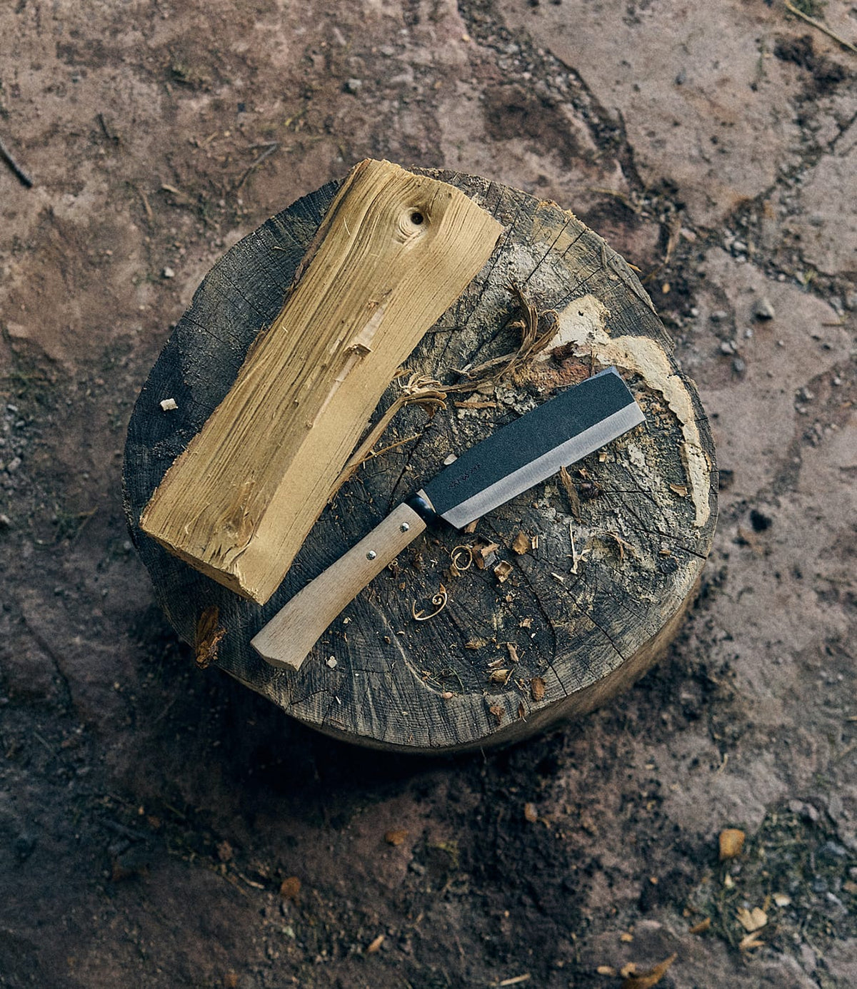 Resting on a weathered tree stump, the Best Made Co. Sayanata Hatchet—with its versatile carbon steel blade—lies beside a piece of split wood, set against a backdrop of stone or dirt ground.