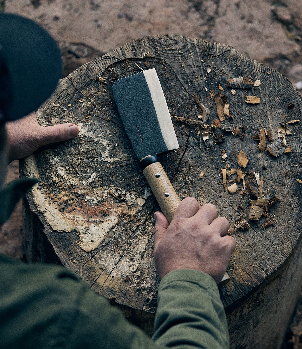 A person holding a compact Best Made Co. Azuma Hatchet with a wooden handle over a worn tree stump, surrounded by wood chips and bark, embodies the rugged functionality of this high-carbon steel tool.
