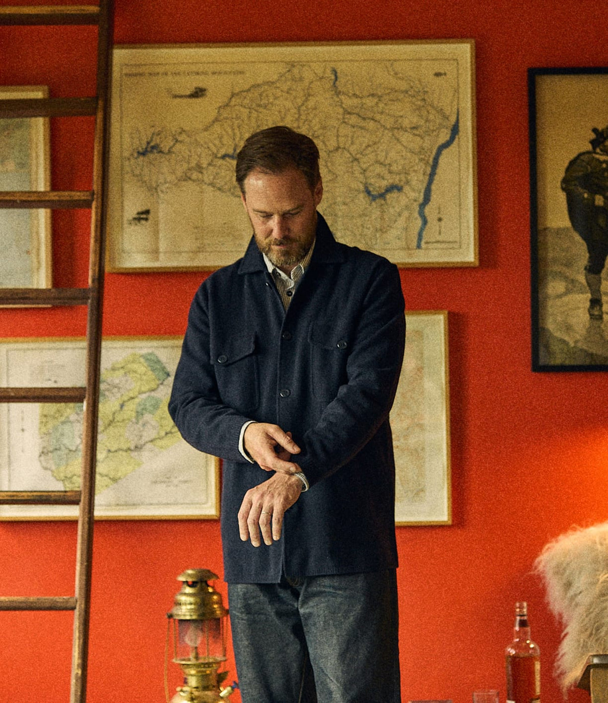 A man wearing a dark Knit Lambswool CPO adjusts his cuff while standing in front of a red wall decorated with maps and framed art.