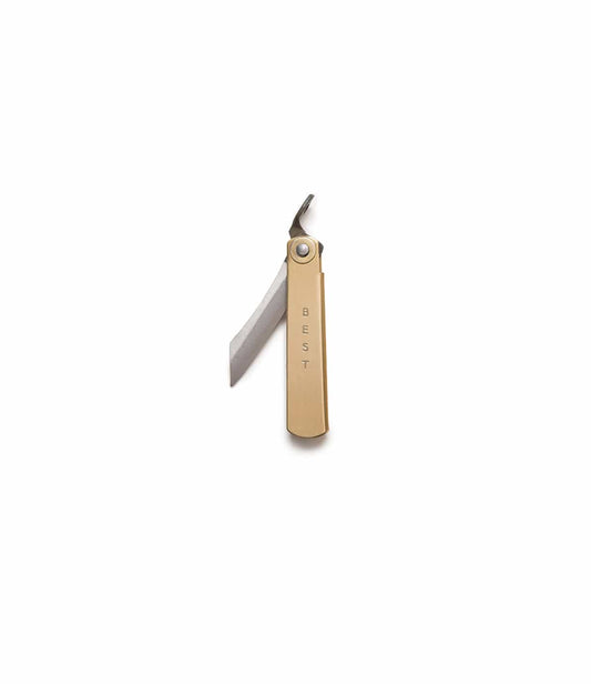 A hand holds a Mini Brass Higo Knife by Best Made Co on a stone sharpening block, with other tools partly visible on a green cutting mat.