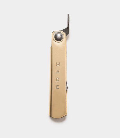 The Mini Brass Higo Knife by Best Made Co. is a gold-colored bottle opener inspired by Japanese folding knives, featuring "MADE" engraved vertically on its handle and shown against a light, plain background.