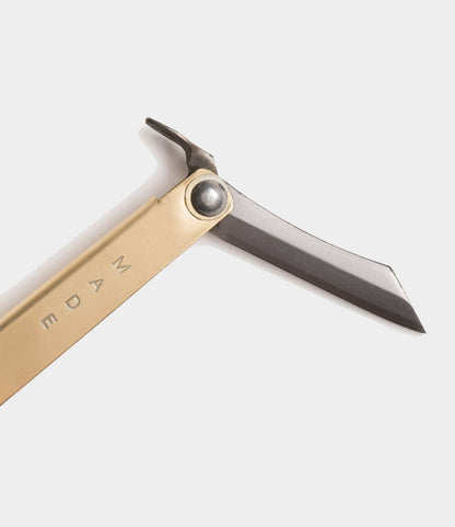 The Best Made Co. Mini Brass Higo Knife, inspired by Edo-period design, displays "MADE" engraved on its brass handle and features a modern black blade with a minimalist look against a light background.