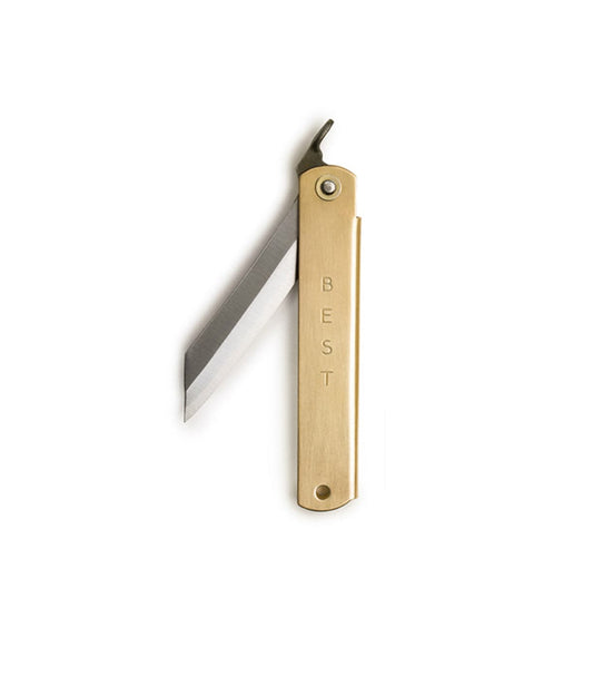 A hand holds the Max Brass Higo Knife by Best Made Co, engraved with MUST, resting on a sharpening stone. Nearby are a Best Made Co box and a sharpening tool on a green cutting mat.