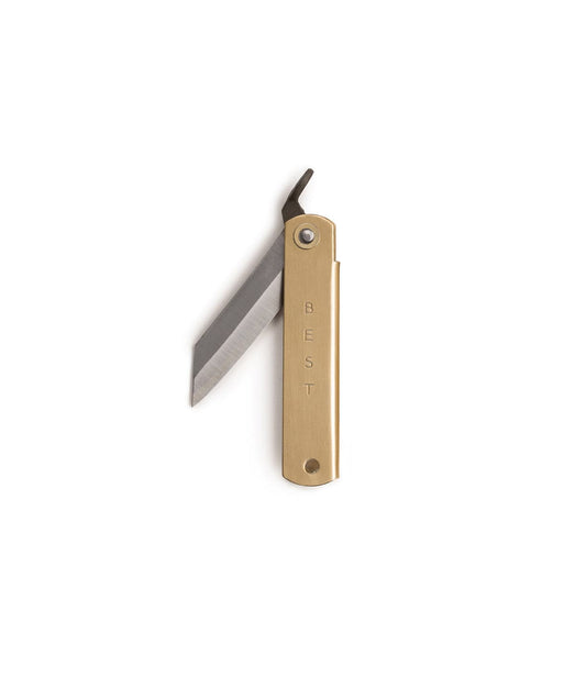A hand holds a partially open Mid Brass Higo Knife from Best Made Co on a sharpening stone beside its branded box and a metal tool, all resting on a green cutting mat.