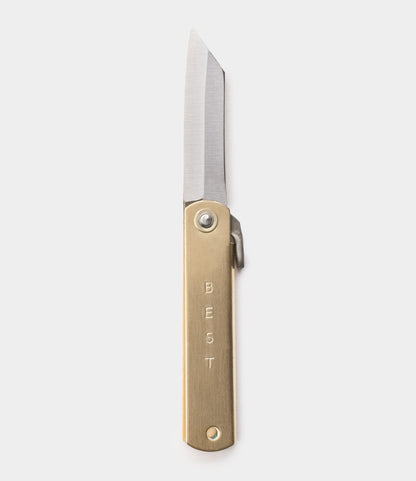The Mid Brass Higo Knife by Best Made Co. features a straight-edged blade and a brass handle engraved vertically with "BEST." It is shown against a plain, light gray background.
