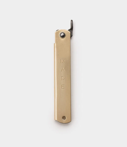 The Max Brass Higo Knife by Best Made Co. is a gold-colored brass bottle opener with "MADE" engraved vertically on its handle, inspired by Japanese folding knives and displayed against a plain white background.