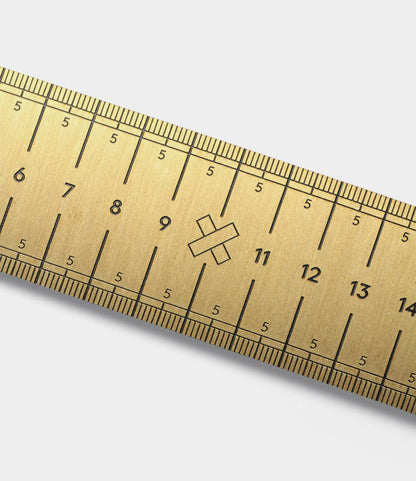 A close-up of the Measure Twice 12 inch Solid Brass Ruler by Best Made Co. shows precise centimeter markings and a decorative X near the 10 cm mark on its brushed brass surface. Made in the USA.