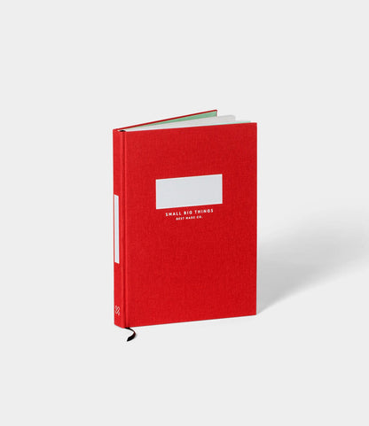 A red S.B.T. Mid Cloth-Bound Notebook by Best Made Co. stands upright on a white background, featuring a white rectangular front label and a black ribbon bookmark hanging from the bottom of its hardcover.