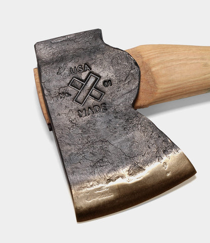 A close-up of the Old Gold No. 1 axe by Best Made Co., featuring a hand-forged, high-carbon steel head with a rough texture, polished blade edge, and USA MADE plus cross icon stamp, set on a wooden handle.