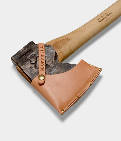The Old Gold No. 1 by Best Made Co. features a hand-forged axe with a wooden handle, metal head stamped USA, and a tan leather blade cover with brass rivets and a BE OPTIMISTIC tag.