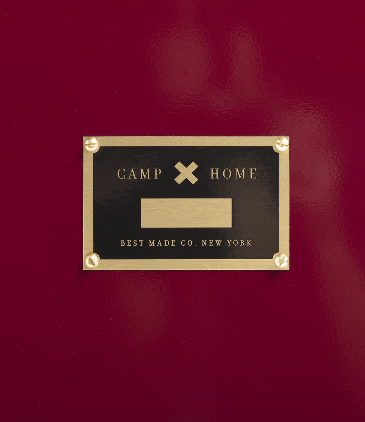 A gold and black plaque on a red surface reads, “CAMP x HOME, BEST MADE COMPANY ARCHIVE, NEW YORK,” featuring a blank area in the center—perfect for labeling your Rolling Footlocker, crafted from premium 22-gauge steel.