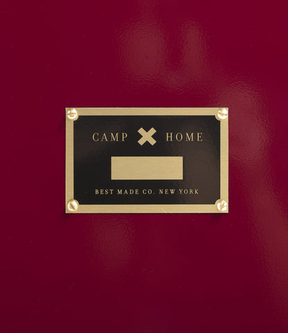 A gold and black plaque on a red surface reads, “CAMP x HOME, BEST MADE COMPANY ARCHIVE, NEW YORK,” featuring a blank area in the center—perfect for labeling your Rolling Footlocker, crafted from premium 22-gauge steel.